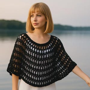 May include: A black, open-weave crochet poncho with a rounded neckline. The poncho is worn over a white garment. The background features a body of water and a blurred treeline.