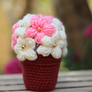 May include: A crocheted flower pot with pink and white flowers. The pot is a deep red colour and the flowers have gold beads in the centre.