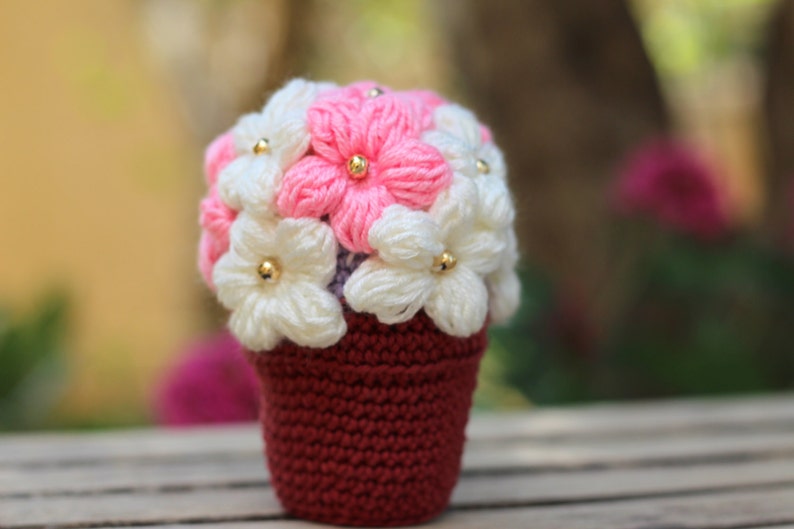 May include: A crocheted flower pot with pink and white flowers. The pot is a deep red colour and the flowers have gold beads in the centre.
