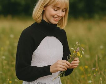 May include: A black knit bolero with a high turtleneck and long sleeves worn over a white sleeveless top featuring a delicate lace overlay with a floral pattern and a scalloped hemline. The garment combination provides a layered appearance.