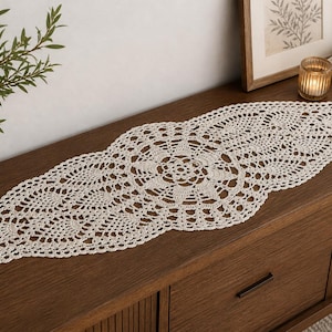 May include: Cream-coloured, oval-shaped crochet table runner. The runner features a central floral design and an openwork pattern. It is displayed on a dark wood surface, alongside a vase with greenery, a framed picture, and a candle.