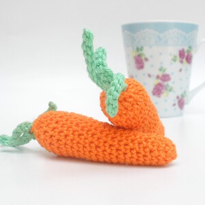 May include: Two crocheted carrots with green tops. The carrots are orange and the tops are a light green. The carrots are lying on a white surface.