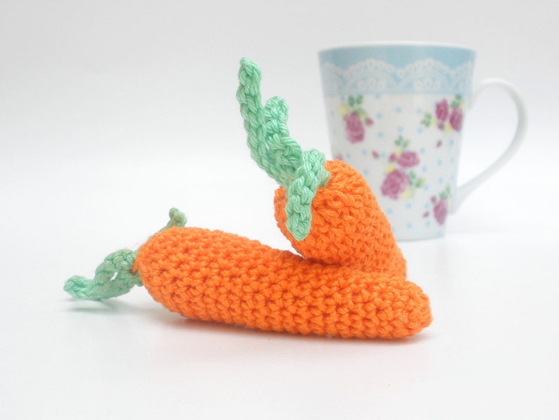 May include: Two crocheted carrots with green tops. The carrots are orange and the tops are a light green. The carrots are lying on a white surface.