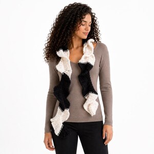 Puede incluir: Bufanda negra y crema con un diseño ondulado. La bufanda está hecha de un material suave y texturizado y tiene detalles de costuras blancas en los bordes. La bufanda se lleva alrededor del cuello.