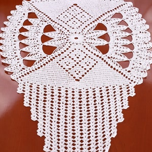 Op de afbeelding: Witte gehaakte tafelloper met een delicaat bloemenpatroon. De loper is gemaakt van fijn garen en heeft een gegolfde rand.