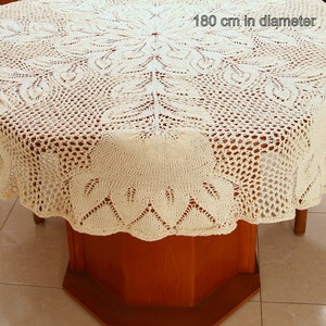 Puede incluir: Un mantel de crochet blanco con un delicado patr&oacute;n floral. El mantel tiene 180 cm de di&aacute;metro y est&aacute; puesto sobre una mesa de madera.