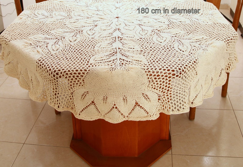 Puede incluir: Un mantel de crochet blanco con un delicado patr&oacute;n floral. El mantel tiene 180 cm de di&aacute;metro y est&aacute; puesto sobre una mesa de madera.