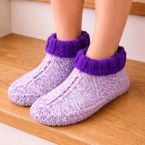 Könnte beinhalten: Ein Paar gestrickte Hausschuhe in Lavendel und Weiß mit einem tiefvioletten Rippbündchen. Die Socken sind handgefertigt und wirken weich und warm, ideal für den Innenbereich. Gezeigt auf einer Holzoberfläche.