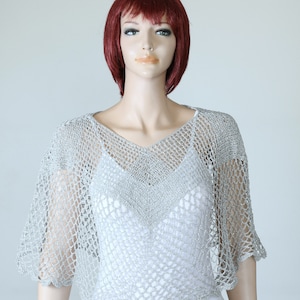 Könnte beinhalten: Eine silberne Häkel-Schal mit einem gewellten Saum. Die Schal ist aus einem zarten, offen gewebten Stoff und hat einen V-Ausschnitt.