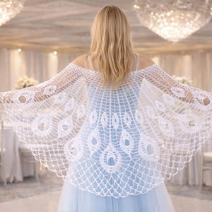 Può includere: Uno scialle all'uncinetto bianco, con spalle scoperte, con un motivo a piume di pavone. Lo scialle è indossato sopra un vestito azzurro. L'intricato motivo traforato è visibile. Lo sfondo presenta una stanza ben illuminata con lampadari e composizioni floreali.