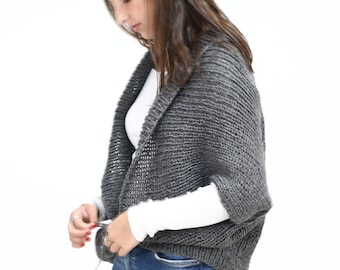May include: A dark gray, chunky knit shrug with a textured, ribbed pattern and short sleeves. The garment features an open front design with a wide collar, crafted from a thick yarn that provides a visible, consistent knit structure throughout.