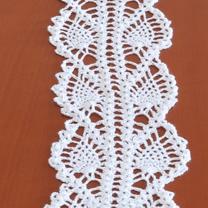 May include: White crocheted table runner with a delicate, lacy design. The runner features a repeating pattern of small, intricate flowers.