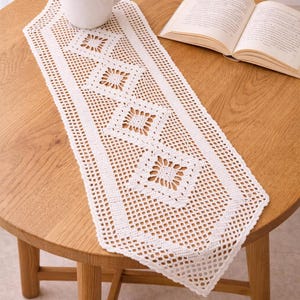 May include: A white, crocheted table runner with a geometric diamond pattern, placed on a round wooden table. A white mug filled with coffee and an open book are also on the table. The runner adds a touch of elegance.