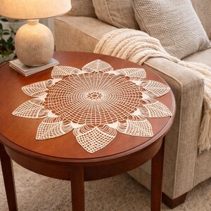 May include: A cream-coloured, crocheted doily with a floral design rests on a round, brown wooden table. The doily has a central circular pattern with radiating petals. A lamp and sofa are in the background.