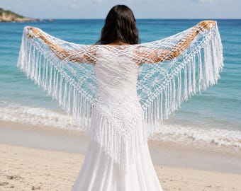 May include: A white triangular crochet shawl featuring an intricate openwork pattern and long fringe along the edges. The shawl is draped across the shoulders, showcasing the detailed yarn work and the delicate, knotted texture of the handmade design.