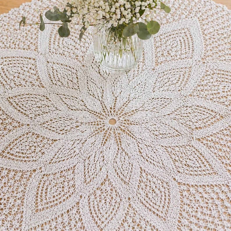 May include: A white, intricately crocheted tablecloth with a floral pattern. The circular tablecloth has a central opening. A glass vase with white flowers and greenery sits on the table, adding a touch of elegance to the scene.