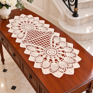 May include: An elongated, off-white crochet table runner featuring a complex, repeating circular motif with a scalloped edge. The intricate needlework displays a central geometric pattern surrounded by fan-like shapes, creating a delicate, openwork texture throughout the entire piece.
