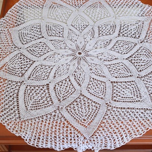 May include: A white, circular, crocheted doily with a floral design. The intricate lace pattern features a central flower shape surrounded by geometric and leaf-like motifs. The doily is displayed on a wooden surface.