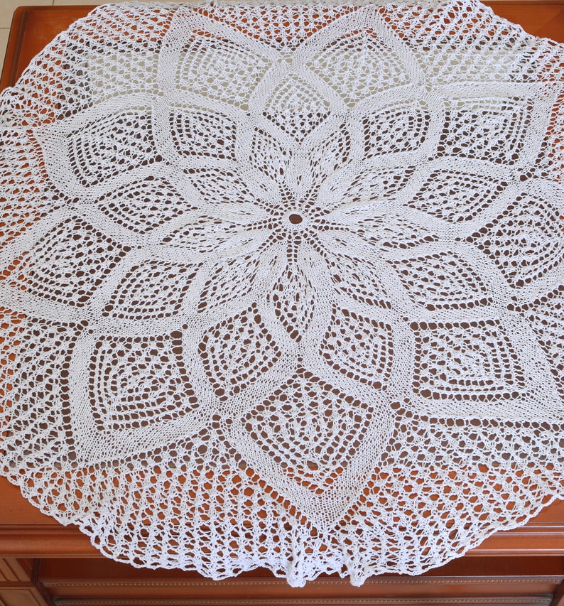 May include: A white, circular, crocheted doily with a floral design. The intricate lace pattern features a central flower shape surrounded by geometric and leaf-like motifs. The doily is displayed on a wooden surface.