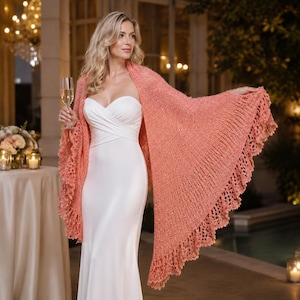 May include: A white strapless floor-length gown paired with a coral-colored knitted shawl. The shawl features a textured, open-knit body and a wide, intricate crocheted lace border along the edges, draped over the shoulders and arms.