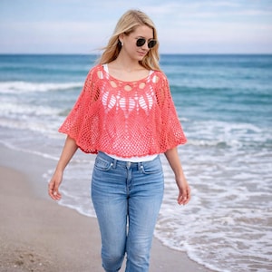 May include: A coral-colored, open-weave crochet poncho with a round neckline and flared sleeves. The poncho is worn over a white top and paired with blue jeans. The background shows a beach and ocean.