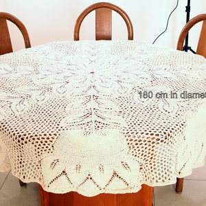 Puede incluir: Un mantel de crochet blanco con un delicado patr&oacute;n floral. El mantel tiene 180 cm de di&aacute;metro y est&aacute; colocado sobre una mesa de madera.