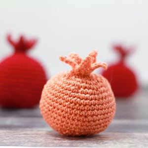 K&ouml;nnte beinhalten: Drei geh&auml;kelte Granat&auml;pfel, einer in Pfirsichfarbe und zwei in Rot. Der Pfirsichgranatapfel ist scharf gestellt, die roten Granat&auml;pfel sind unscharf.