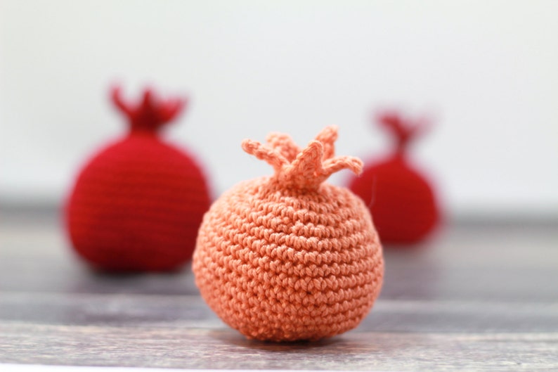 K&ouml;nnte beinhalten: Drei geh&auml;kelte Granat&auml;pfel, einer in Pfirsichfarbe und zwei in Rot. Der Pfirsichgranatapfel ist scharf gestellt, die roten Granat&auml;pfel sind unscharf.