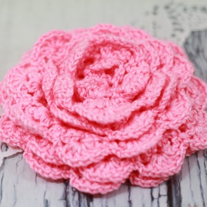 May include: A pink crocheted flower with multiple layers. The flower is made with a delicate stitch pattern and has a soft, feminine look.