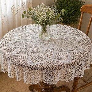May include: A cream-coloured, round crochet tablecloth with a floral pattern covers a wooden table. A glass vase with white flowers sits in the centre. A wooden chair is visible on the right.