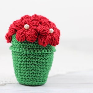 May include: A crocheted flower pot with red flowers and white pearl accents. The pot is green and the flowers are made of yarn.
