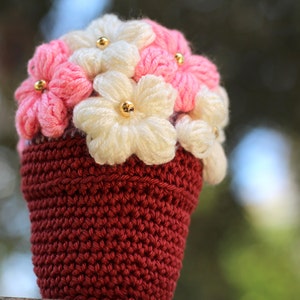 May include: A crocheted flower pot with pink and white flowers. The pot is a deep red colour and the flowers have gold beads in the centre.