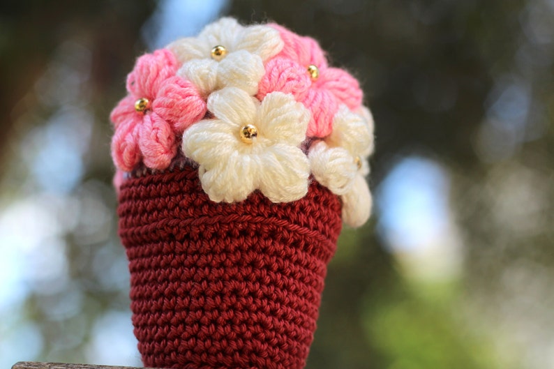 May include: A crocheted flower pot with pink and white flowers. The pot is a deep red colour and the flowers have gold beads in the centre.