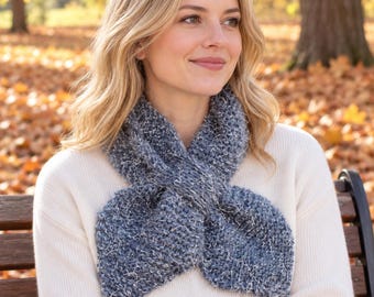 May include: A textured, dark blue knitted scarf with a subtle metallic silver shimmer, featuring a cross-over design. The scarf is worn over a plain, long-sleeved cream-coloured jumper, showcasing a chunky, hand-knit appearance with visible yarn loops.