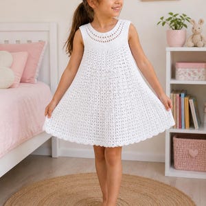 Può includere: Un vestito bianco all'uncinetto senza maniche, con scollo tondo e gonna svasata. L'abito ha un orlo smerlato. La ragazza indossa l'abito e si trova su un tappeto rotondo intrecciato.