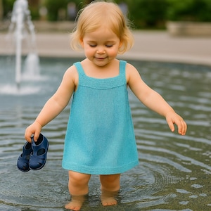 Könnte beinhalten: Ein junges Kind in einem türkisblauen Strickkleid steht im Wasser und hält ein Paar marineblaue Sandalen. Das Kleid hat einen quadratischen Ausschnitt und breite Träger. Das Kind ist barfuß, das Wasser reicht bis zu den Knien.