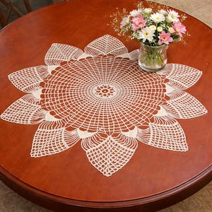 May include: Cream-colored, crocheted doily in a floral design, placed on a round, wooden table. The doily has a central circular pattern with radiating lines and petal-shaped edges. A glass vase with pink and white flowers sits on the table.