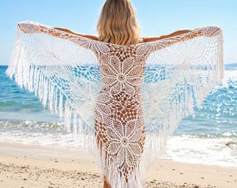 May include: A white crochet shawl featuring an intricate floral pattern with a wide, open-weave design. The triangular garment is finished with long, delicate fringe along the edges, creating a textured and lightweight appearance for the accessory.