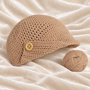 May include: A tan crocheted newsboy cap featuring a textured stitch pattern and a single light-colored button on the side. Next to the cap sits a small, round ball of matching tan yarn, showcasing the same material used for the headwear construction.