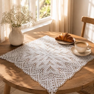 May include: A square white crocheted doily featuring an intricate leaf-like pattern with delicate openwork edges. The doily is centered on a round wooden table, accompanied by a ceramic vase with white flowers, a croissant, and a coffee cup.