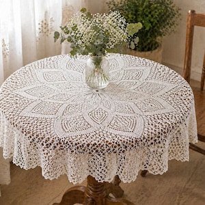 May include: A round, cream-colored lace tablecloth with a floral design covers a wooden table. A clear glass vase with white flowers sits in the center. A wooden chair is to the right.