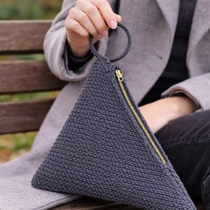 May include: A triangular, dark gray knitted clutch with a circular handle. The clutch features a gold-colored zipper closure along one side. The bag is held by a person sitting on a wooden bench.