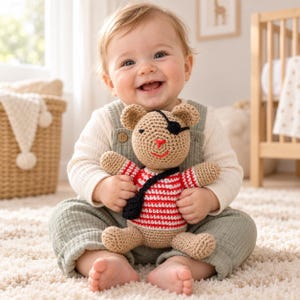 May include: A crocheted tan teddy bear toy featuring a black eye patch over one eye, a red stitched nose and mouth, and a red and white striped shirt. The bear also wears a black crocheted crossbody bag strap across its chest.