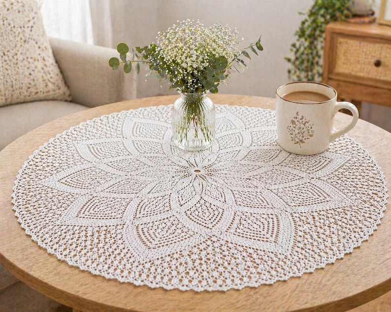 May include: A circular, white, crocheted doily with a detailed floral pattern is on a wooden table. A mug with a floral design and a vase of flowers are also on the table. The doily's intricate design adds a touch of elegance to the setting.