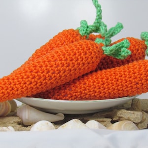 May include: Four crocheted carrots with green tops, arranged on a white plate with seashells on a sandy surface.