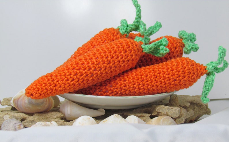 May include: Four crocheted carrots with green tops, arranged on a white plate with seashells on a sandy surface.