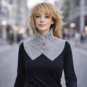May include: A gray knitted cowl with a geometric pattern and a wooden button closure. The cowl is worn over a black long-sleeved shirt. The background is a blurred city street.