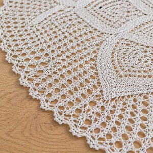 May include: Close-up of a white, crocheted doily with a scalloped edge. The intricate openwork design features geometric patterns and a delicate texture. The doily is placed on a wooden surface.