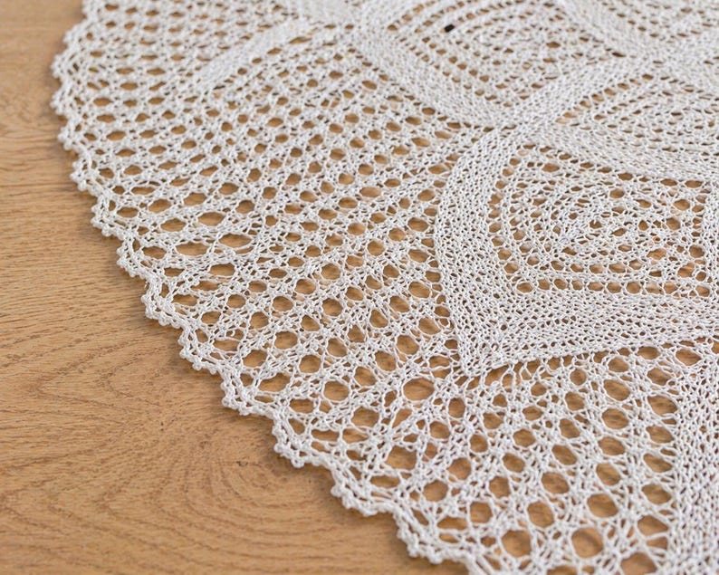 May include: Close-up of a white, crocheted doily with a scalloped edge. The intricate openwork design features geometric patterns and a delicate texture. The doily is placed on a wooden surface.
