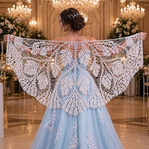 May include: A cream-colored, open-weave crochet shawl with a leaf-like pattern. The shawl is draped over a light blue gown. The background features a grand ballroom with chandeliers and floral arrangements, suggesting a formal event or wedding.